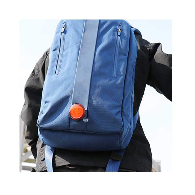 Person wearing a blue backpack with a red Cateye rear safety light clipped to the back, increasing visibility while cycling or walking.